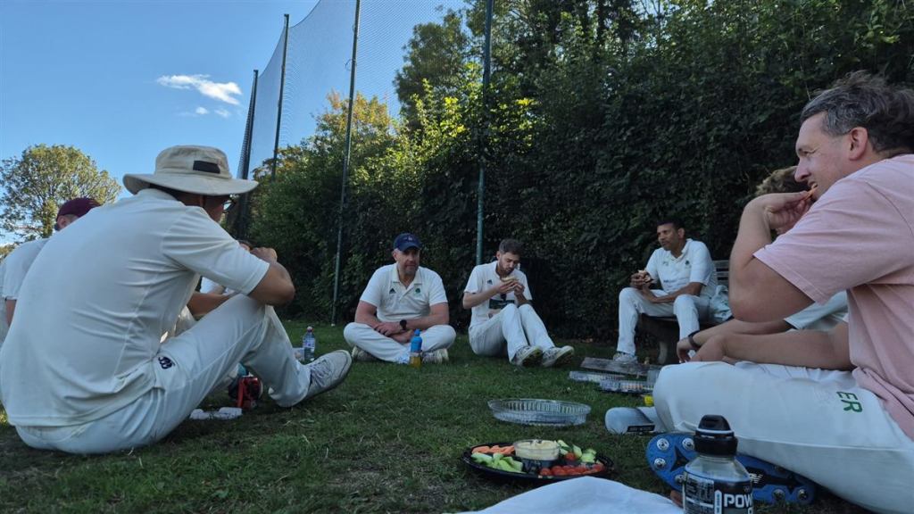 Park Hill Cricket Club players sitting on the grass beside the practice nets at the tea interval, with a platter of carrot and cucumber sticks and hummus